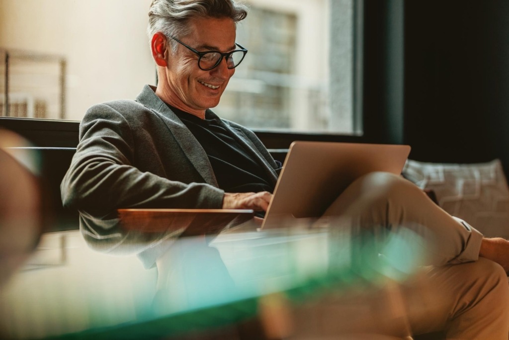 Man smiling while reading on a laptop while sitting on a couch in front of a window